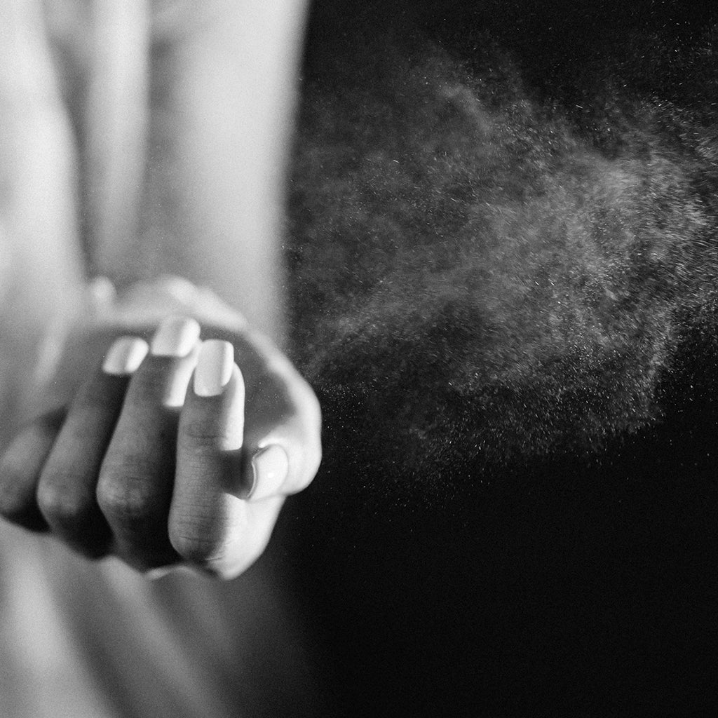 black and white image of a lady spraying perfume across wrist of left arm. A mesmerising cloud of mist, created by the droplets of oud are visible