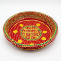 A round, handwoven decorative tray or basket with intricate embroidery. The base is a vibrant red, decorated with yellow central motifs, yellow flower-like accents, and a border of green and yellow geometric patterns, all set against a plain white background.
